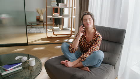 Happy-young-woman-sits-in-modern-living-room-and-talks-on-phone,-wide