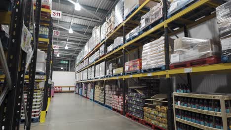 Warehouse-interior-showing-rows-of-high-shelves-stocked-with-various-goods-in-a-wholesale-store,-industrial-lighting