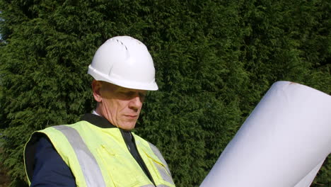 A-construction-manager-architect-standing-in-front-of-trees-looking-at-plans-and-paperwork
