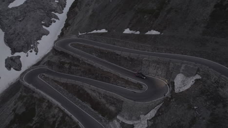 Drone-shot-of-a-car-driving-down-the-hill-on-the-Stelvio-Pass-Italy-on-a-grey-day-with-snow-on-the-mountains-on-a-twisty-road-and-headlights-on-during-sunset-LOG