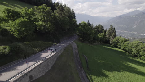 Drone-shot-of-a-lonely-grey-car-driving-in-between-the-trees-and-woods-on-a-sunny-day-in-the-Alps-with-a-panoramic-view-near-Bolzano-Italy-LOG