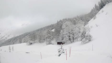 Malerische,-Langsame-Hüttenlandschaft-Inmitten-Verschneiter-Berghänge,-Schneebedeckter-Bäume,-Leichtem-Schneefall-Und-Bewölktem-Himmel