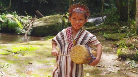 Young-indigenous-boy-smiling-in-traditional-attire-playing-a-drum-in-Pucallpa,-Peru,-lush-forest-backdrop,-daylight