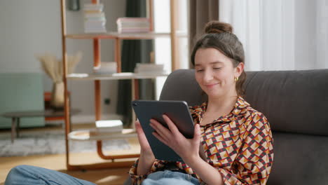 Smiling-young-caucasian-woman-sits-in-living-room-and-uses-tablet