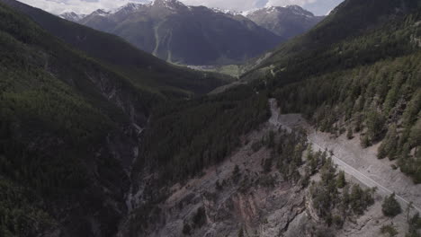 Drone-shot-flying-near-the-Fluela-Pass-Italy-on-a-sunny-and-cloudy-day-revealing-the-snowy-mountains-in-the-background-and-a-road-on-the-right-side-of-the-scene-in-between-forests-and-trees-LOG