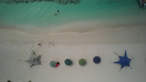 Vibrant-umbrellas-and-tourists-on-the-white-sands-of-los-roques,-venezuela,-turquoise-water,-aerial-view