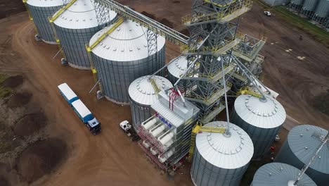 Industrial-grain-silos-with-trucks-loading,-overcast-day,-aerial-view