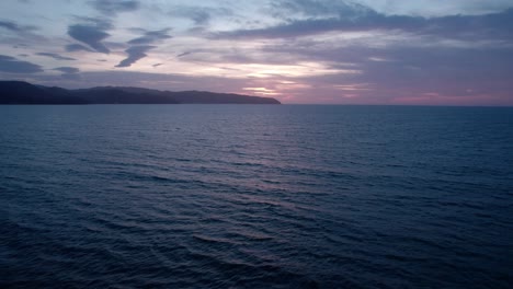 Twilight-hues-over-calm-sea-with-distant-mountains-under-a-serene-sky,-aerial-view