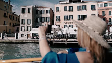 Caucasian-women-with-a-hat-and-sunglasses-taking-photos-with-her-phone-during-a-boat-trip-exploring-the-scenic-city-of-Venice-in-italy