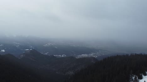 Incredible-aerial-drone-shot-of-mountains-full-of-pine-trees-and-snow,-and-a-foggy-sky-during-a-winter-snowfall