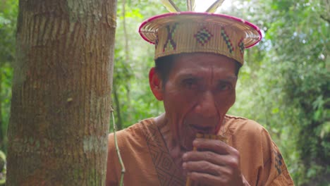 Indigenous-man-in-traditional-attire-playing-flute-in-Pucallpa,-Peru,-surrounded-by-lush-forest