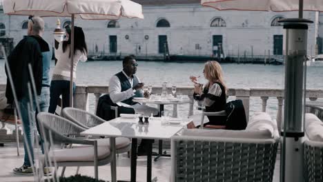 Classy-mixed-race-couple-with-sunglasses-eating-ice-cream-on-a-terrace-at-the-grand-canale-in-Venice-italy-and-talking-while-boats-floating-by-on-a-sunny-day---luxury-jetset-travel-lifestyle