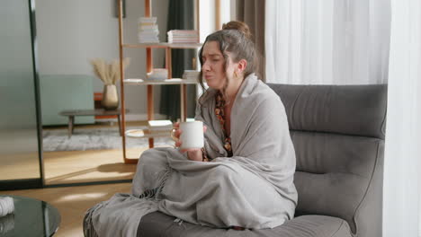 Sick-young-woman-wrapped-in-blanket-drinks-tea-in-modern-living-room
