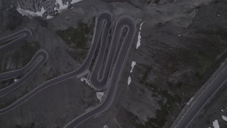 Top-down-drone-shot-of-a-car-driving-down-the-hill-on-the-Stelvio-Pass-Italy-on-a-grey-day-with-snow-on-the-mountains-on-a-twisty-road-and-headlights-on-during-sunset-LOG