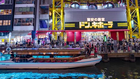 Night-time-lapse-of-people-getting-on-tour-boats-outside-the-Ebisu-Tower-Ferris-Wheel-Don-Quixote-at-the-Dotonbori-Canal-Namba-Osaka,-Japan-TILT