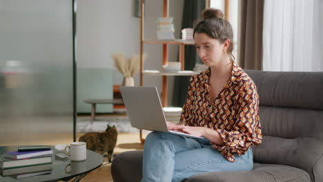 Woman-uses-laptop-in-living-room-while-cat-walks-in-background,-slomo