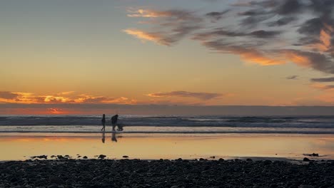 Sunset-timelapse-with-silhouetted-people-and-pets-enjoying-the-beach-low-profile-with-rocks-in-foreground