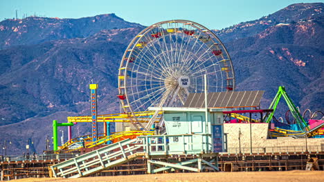 Santa-Monica-Pier-amusement-rides,-beach,-lifeguard-shack,-and-mountains-in-the-background---daytime-time-lapse