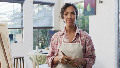 Portrait-of-biracial-female-artist-in-apron-holding-brushes-in-studio,-slow-motion