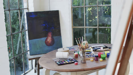 Close-up-of-paintings-on-easels-and-paints-and-brushes-on-table-in-studio,-slow-motion