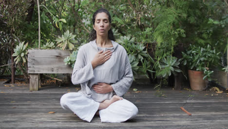 Mujer-Birracial-Enfocada-Practicando-Meditación-De-Yoga-En-Un-Jardín-Soleado,-Cámara-Lenta