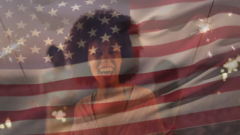 Zusammengesetztes-Video-Der-US-Flagge-über-Einer-Glücklichen-Afroamerikanischen-Frau,-Die-Am-Strand-Mit-Wunderkerze-Feiert