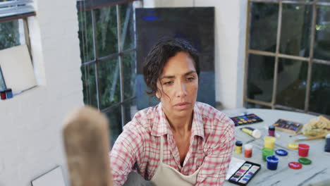 Biracial-female-artist-in-apron-holding-brush-and-looking-at-canvas-in-studio,-slow-motion