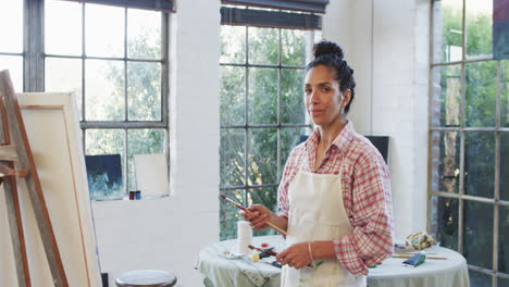 Portrait-of-biracial-female-artist-in-apron-holding-brushes-in-studio,-slow-motion
