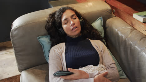 Biracial-woman-lying-on-sofa-under-blanket-listening-music-using-smartphone-at-home,-slow-motion
