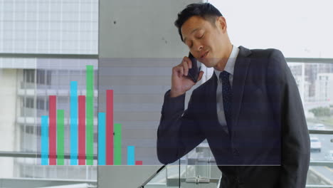 Animation-Von-Mehrfarbigen-Balkendiagrammen-über-Einem-Asiatischen-Mann,-Der-Im-Büro-Auf-Dem-Mobiltelefon-Spricht