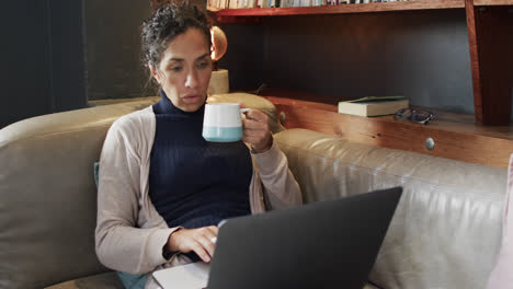 Biracial-woman-sitting-on-sofa-using-laptop-and-drinking-coffee-at-home,-slow-motion