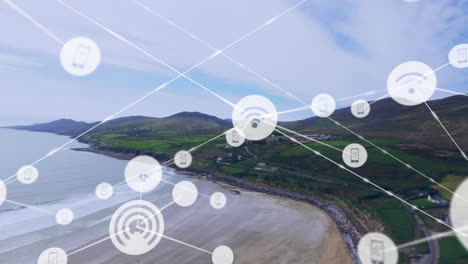 Animation-of-network-of-digital-icons-against-aerial-view-of-landscape-with-mountains,-beach-and-sea