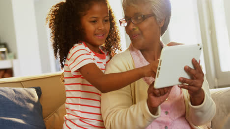 Smiling-grandmother-and-granddaughter-using-digital-tablet-on-sofa-in-living-room-4k
