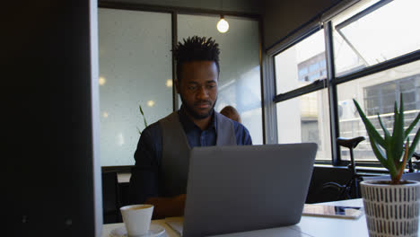 Front-view-of-young-black-businessman-working-on-laptop-with-a-cup-of-coffee-sitting-at-desk-4k