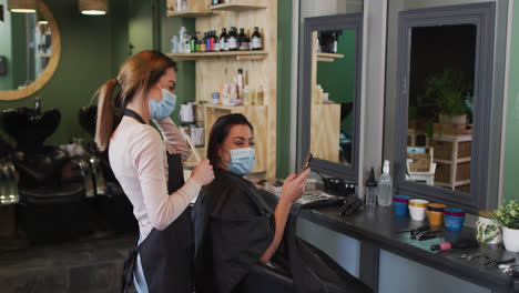 Female-hairdresser-and-female-customer-wearing-face-masks-looking-at-smartphone-at-hair-salon