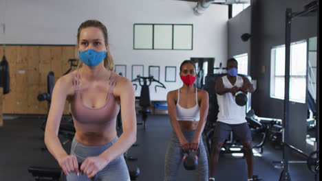 Two-fit-caucasian-women-and-fit-african-american-man-wearing-face-masks-exercising-using-kettlebell-