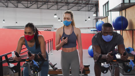 Caucasian-female-fitness-trainer-wearing-face-mask-at-gym-standing-by-stationary-bikes