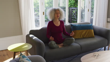 Ältere-Kaukasische-Frau-Praktiziert-Yoga,-Während-Sie-Zu-Hause-Auf-Der-Couch-Sitzt