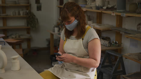 Female-caucasian-potter-wearing-face-mask-and-apron-using-smartphone-at-pottery-studio