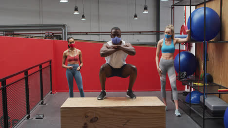 African-american-man-jumping-on-box-wearing-face-mask-at-gym,-two-diverse-women-watching