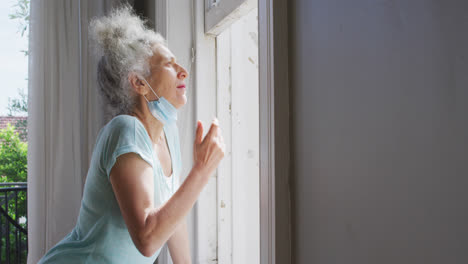 Senior-caucasian-woman-with-lowered-face-mask-looking-out-of-the-window-at-home