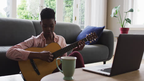 Mujer-Afroamericana-Tocando-La-Guitarra-Mientras-Mira-La-Computadora-Portátil-En-Casa