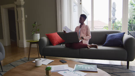 African-american-woman-reading-a-document-and-using-laptop-while-working-from-home