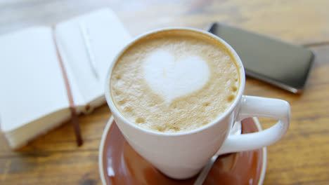 Cup-of-hot-coffee-latte-with-heart-shaped-foam-art-with-diary-and-mobile-phone-4k