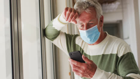 Senior-caucasian-man-wearing-face-mask-using-smartphone-near-window-at-home