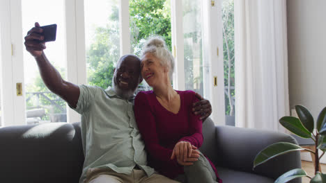 Mixed-race-senior-couple-taking-a-selfie-with-smartphone-in-the-living-room-at-home