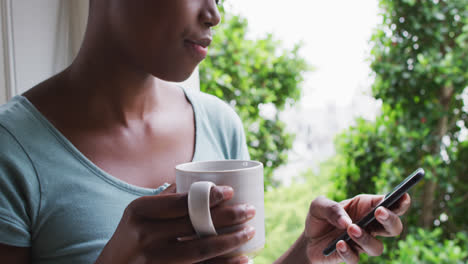 Afroamerikanische-Frau-Hält-Kaffeetasse-Mit-Smartphone-Zu-Hause