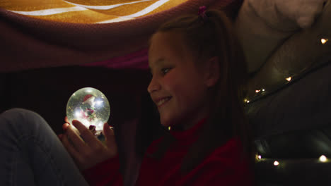 Caucasian-girl-smiling-while-holding-snow-globe-under-blanket-fort-during-christmas-at-home