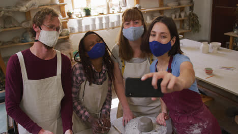 Diverse-potters-wearing-face-masks-and-aprons-taking-a-selfie-using-smartphone-at-pottery-studio