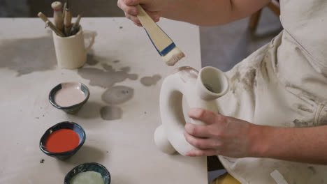 Mid-section-of-female-potter-wearing-apron-using-glaze-brush-to-paint-on-pot-at-pottery-studio